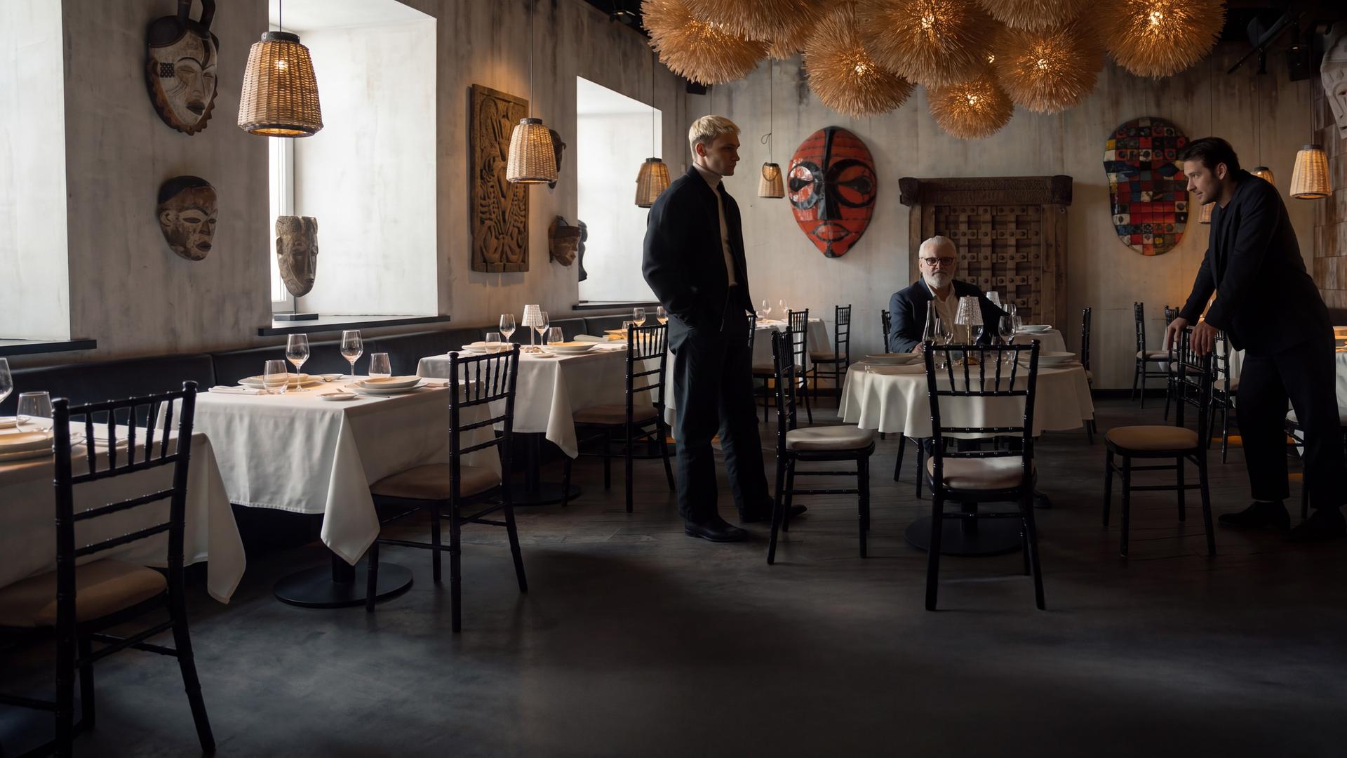 "Serial Cleaner - Absolute Stille": Ludwig Karanz (László I. Kish) sitzt am Tisch im Restaurant. Seine beiden Söhne Christian Karanz (Rick Okon) und Vincent Karanz (Stefan Gorski) stehen sich davor angespannt gegenüber.