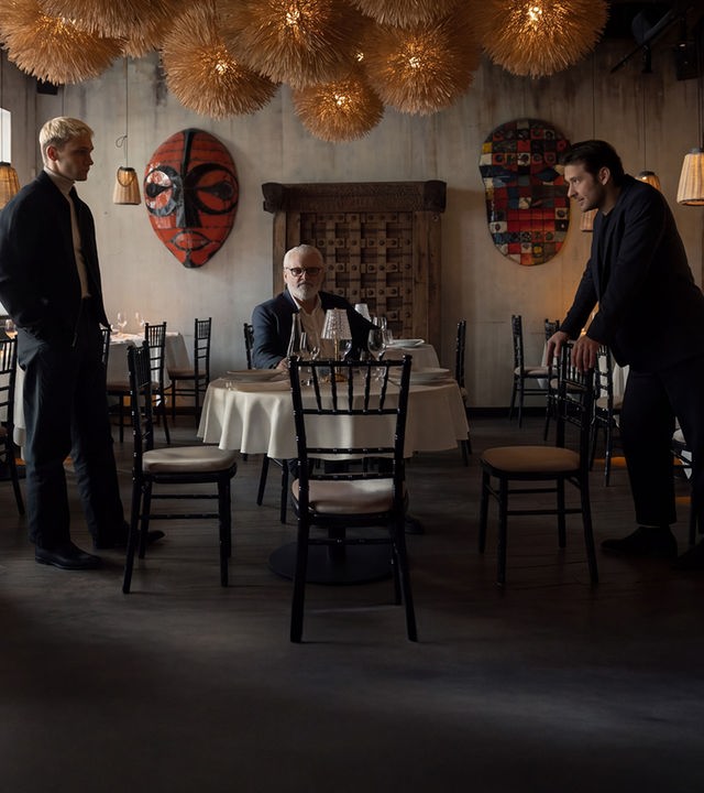 "Serial Cleaner - Absolute Stille": Ludwig Karanz (László I. Kish) sitzt am Tisch im Restaurant. Seine beiden Söhne Christian Karanz (Rick Okon) und Vincent Karanz (Stefan Gorski) stehen sich davor angespannt gegenüber.