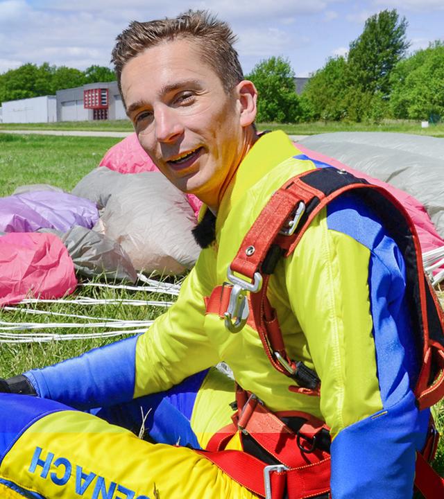 Moderatror Eric Mayer sitzt auf einer Wiese am Boden. Um ihn herum liegt ein Fallschirm. Er hat einen Schutzanzug an und darüber trägt er das Geschirr, dass ihn mit dem Co-Tandem-Fallschirmspringer und dem Fallschirm verbunden hat. Rechts hinter ihm kniet der Fallschirmspringer, der Eric beim Tandemsprung mitgenommen hat.