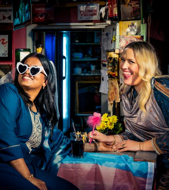 Hinduistin Sagitha blickt lächelnd seitlich nach vorne. Sie trägt eine Sonnenbrille in Herzform, auf der "just married" steht, und einen Schleier. Atheistin Josi sitzt im Sari neben ihr und streckt ihre Zunge heraus.