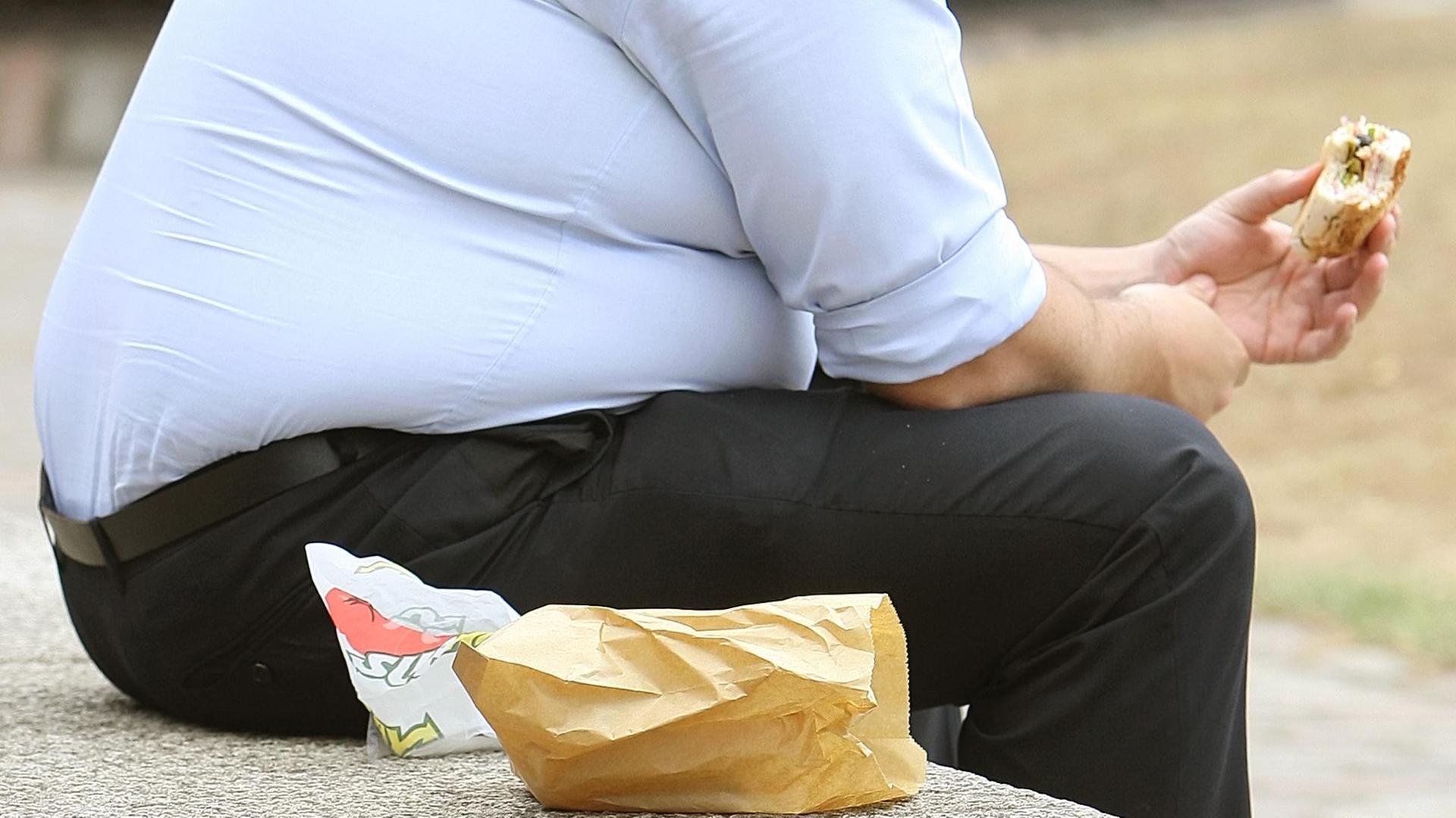 Übergewichtiger Mann sitzt in einem Park und isst einen Hamburger