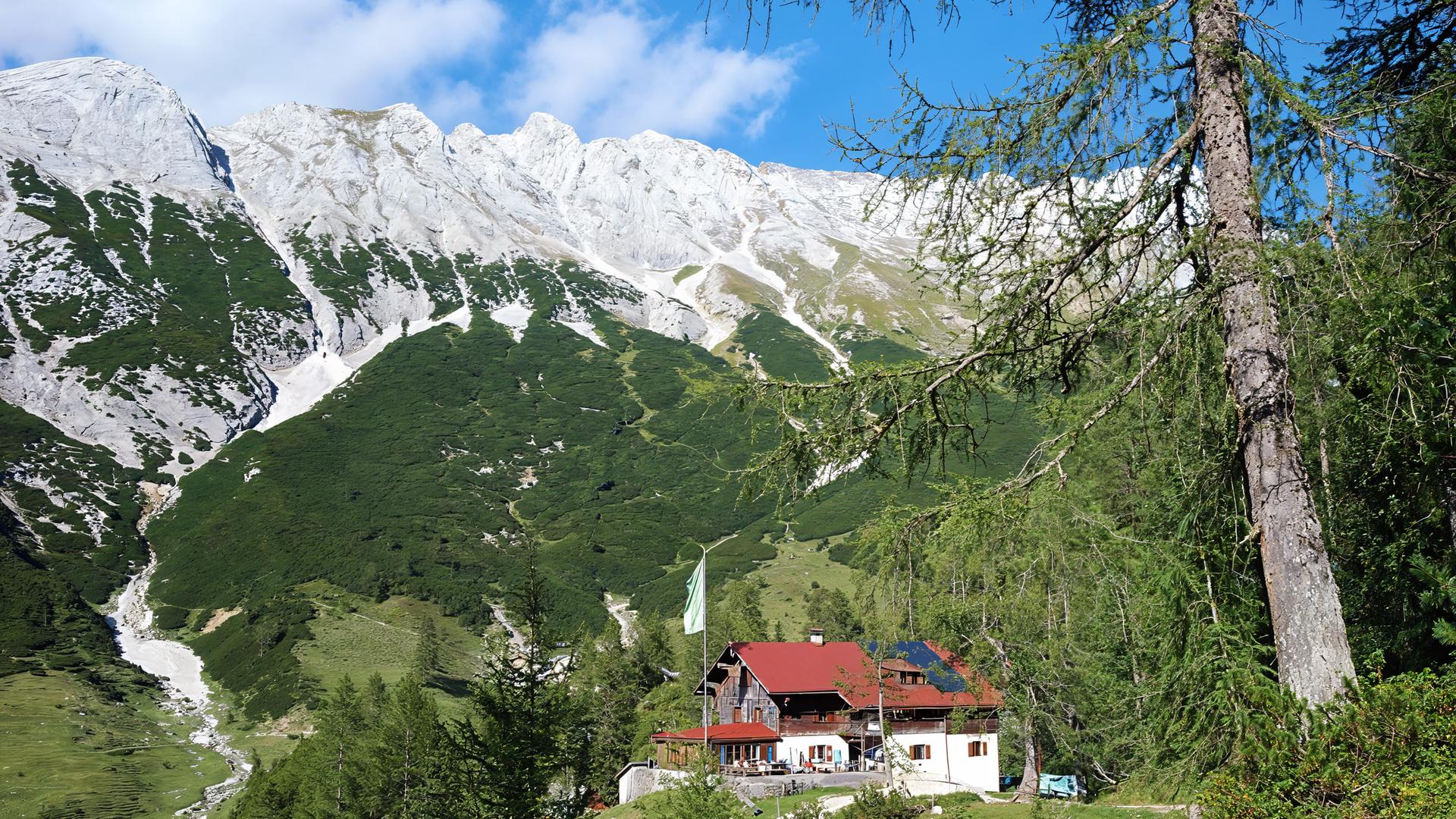 Eine Alpenvereinshütte mit rotem Dach liegt eingebettet in grüne Almwiesen und Nadelwälder vor der imposanten Kalkfelskulisse des Karwendelgebirges.