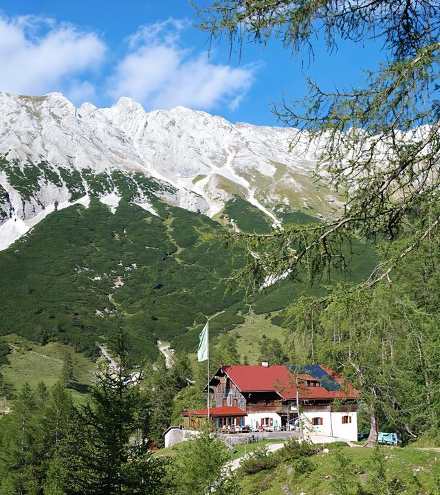 Eine Alpenvereinshütte mit rotem Dach liegt eingebettet in grüne Almwiesen und Nadelwälder vor der imposanten Kalkfelskulisse des Karwendelgebirges.