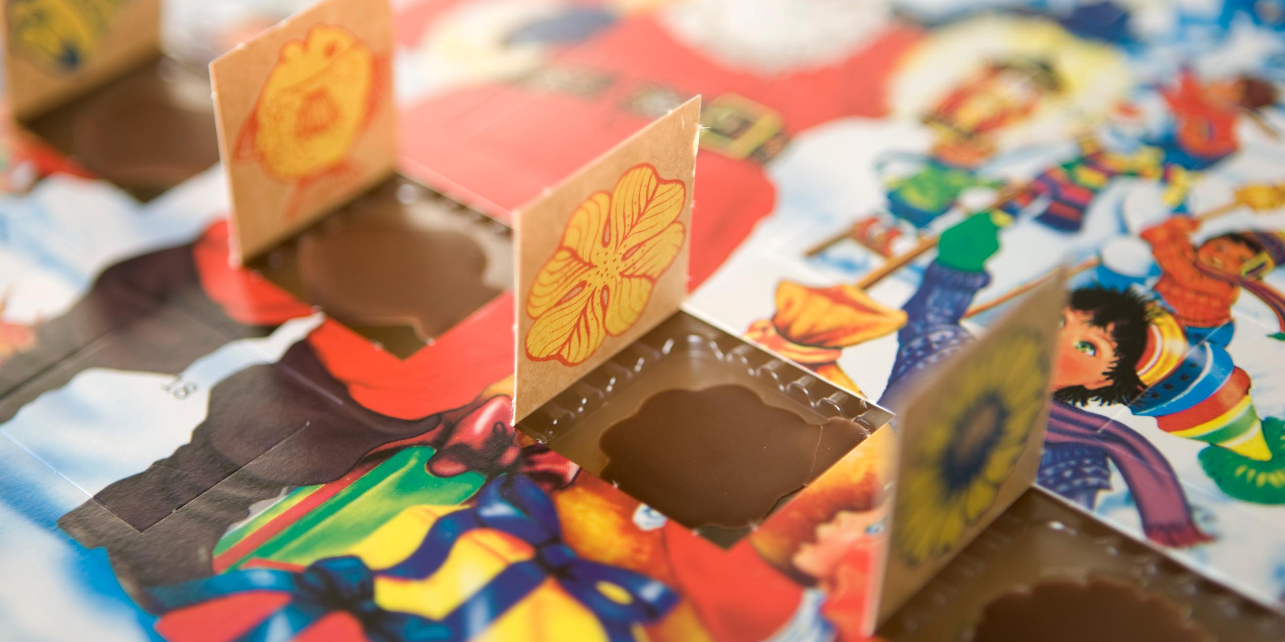 Man sieht einen Adventskalender mit winterlichem Motiv an dem vier Türchen nebeneinander geöffnet sind. Die kleinen Schokoladenmotive liegen noch in ihren Förmchen. 