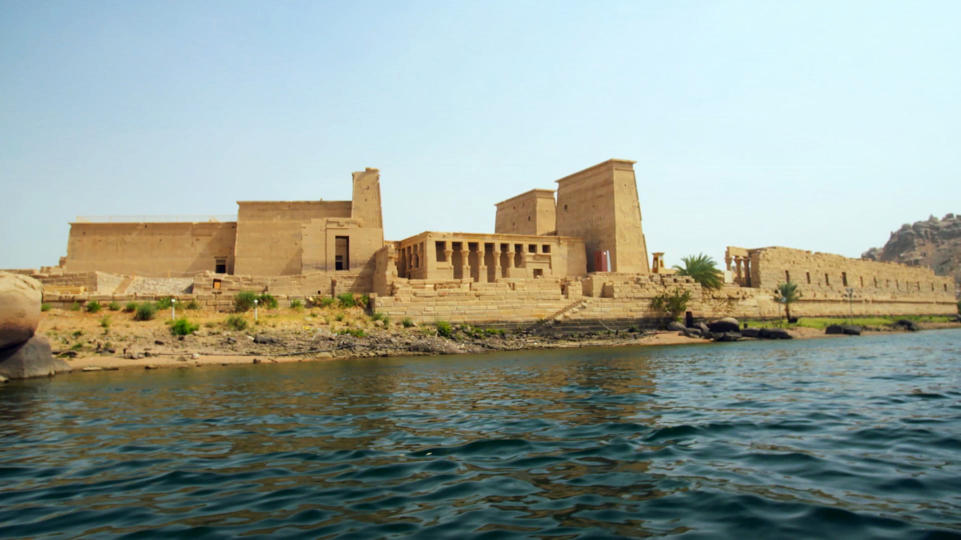 Ein großer, sandsteinfarbener Tempelkomplex mit mehreren Gebäuden ragt am Ufer eines Flusses empor, der im Vordergrund zu sehen ist. Der mittlere, zwischen zwei großen Doppelpylonen gelegene Teil des Tempels weist mehrere Säulen auf. An der Seite stehen weitere Gebäude der Tempelanlage, darunter ebenfalls ein Fragment mit mehreren Säulen. Am Ufer des Flusses stehen zwei Palmen. Grüne, niedrige Pflanzen wachsen zwischen Geröll. Der Himmel ist strahlendblau.