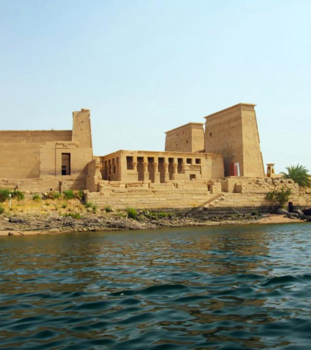 Ein großer, sandsteinfarbener Tempelkomplex mit mehreren Gebäuden ragt am Ufer eines Flusses empor, der im Vordergrund zu sehen ist. Der mittlere, zwischen zwei großen Doppelpylonen gelegene Teil des Tempels weist mehrere Säulen auf. An der Seite stehen weitere Gebäude der Tempelanlage, darunter ebenfalls ein Fragment mit mehreren Säulen. Am Ufer des Flusses stehen zwei Palmen. Grüne, niedrige Pflanzen wachsen zwischen Geröll. Der Himmel ist strahlendblau.