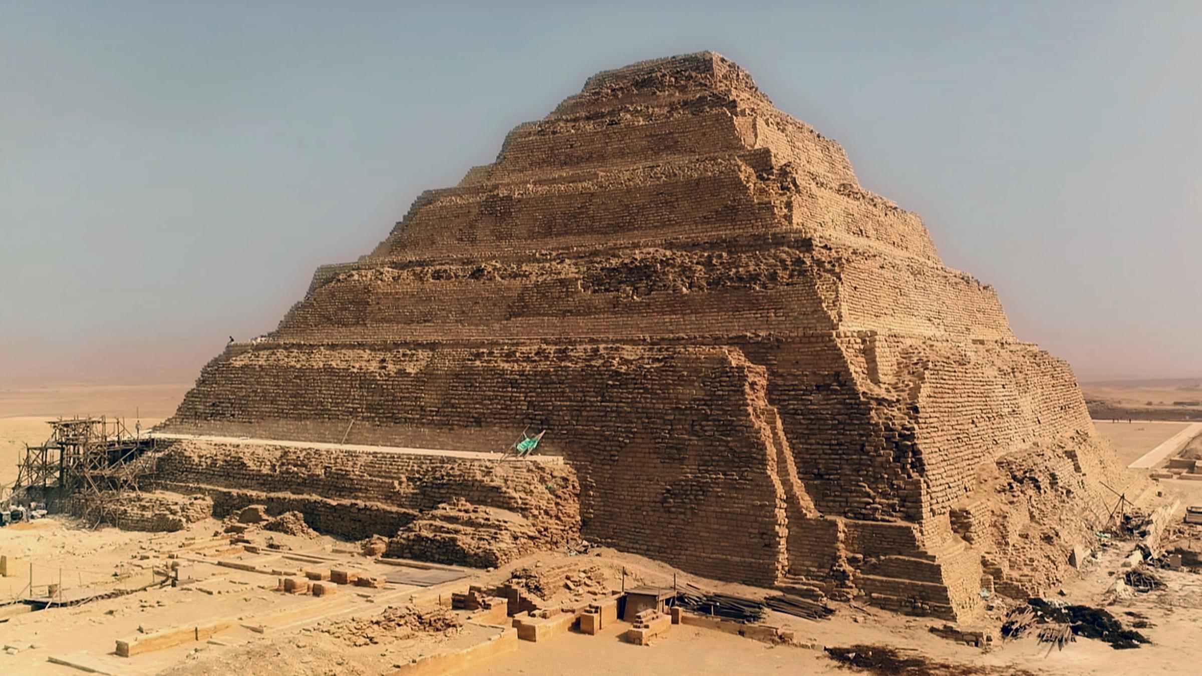 Totale der Djoser-Pyramide mit ihren Steinstufen. Die Verkleidung ist teilweise zerstört. Im Hintergrund erheben sich mehrere kleinere Bauten und Strukturen.