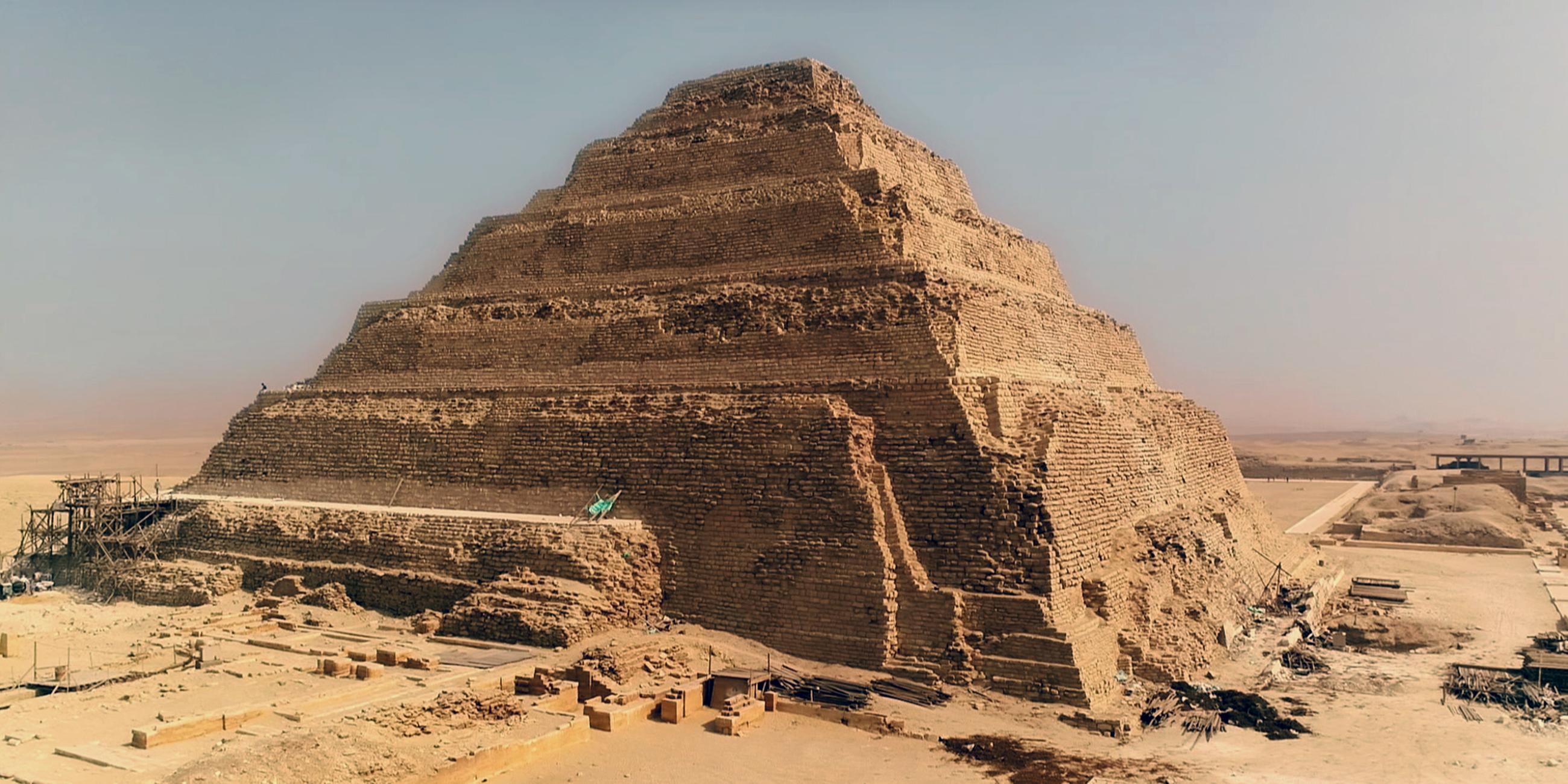 Totale der Djoser-Pyramide mit ihren Steinstufen. Die Verkleidung ist teilweise zerstört. Im Hintergrund erheben sich mehrere kleinere Bauten und Strukturen.