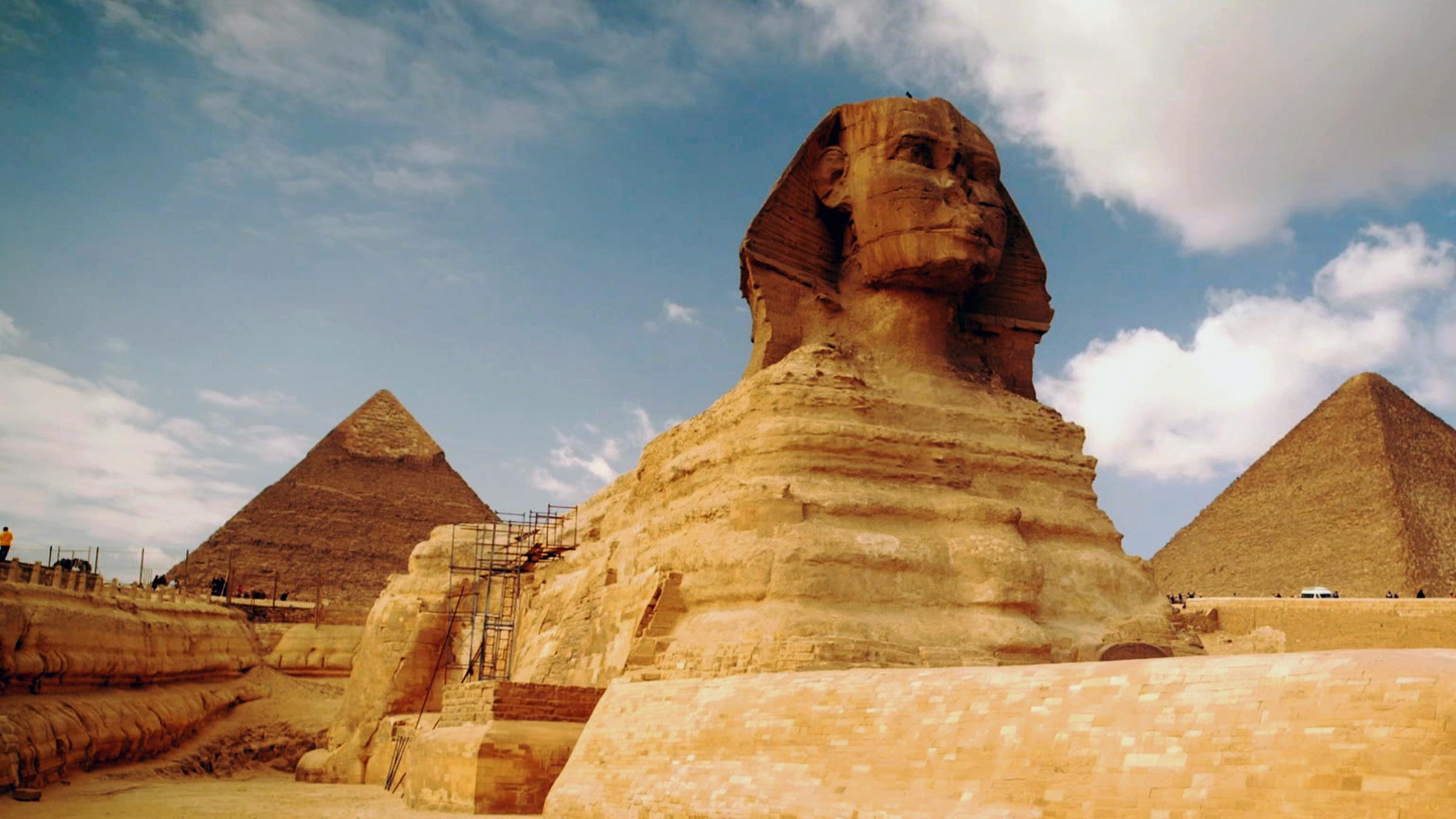 Schrägansicht von unten auf die große Sphinx von Gizeh. An der linken Seite der Sphinx steht ein Baugerüst. Der Himmel ist bewölkt. Im Hintergrund sind zwei Pyramiden zu erkennen.