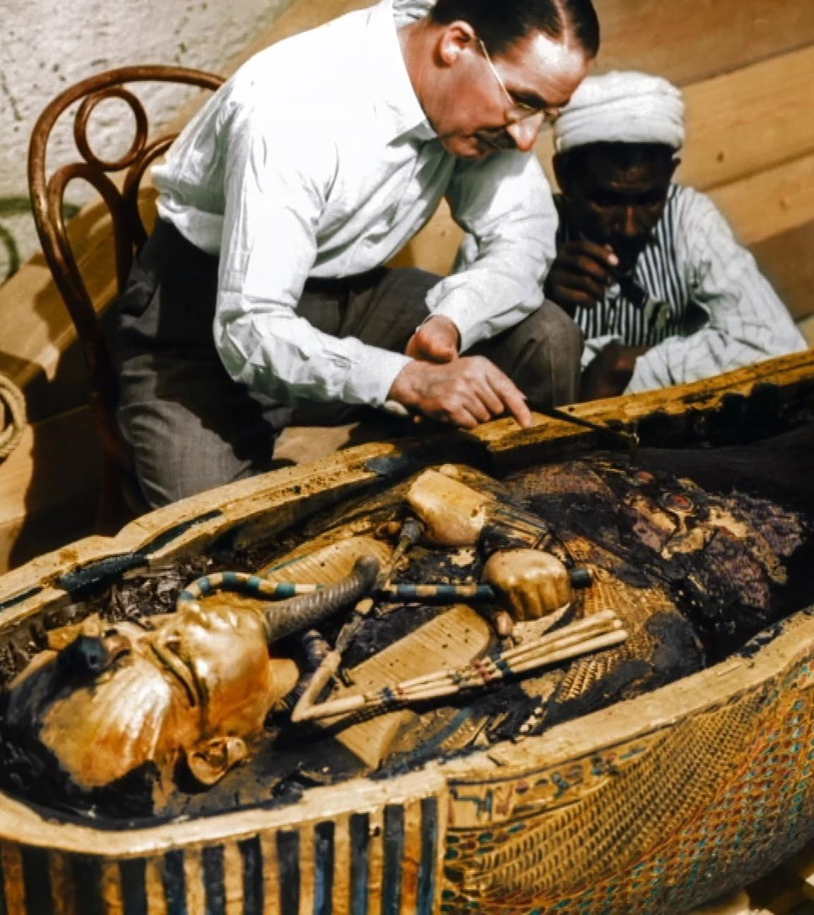 Howard Carter kniet in einer kleinen Kammer neben einem geöffneten Sarkophag. Darin liegt eine Mumie, bedeckt mit einer goldenen Maske. Neben ihm kniet ein weiterer Mann.