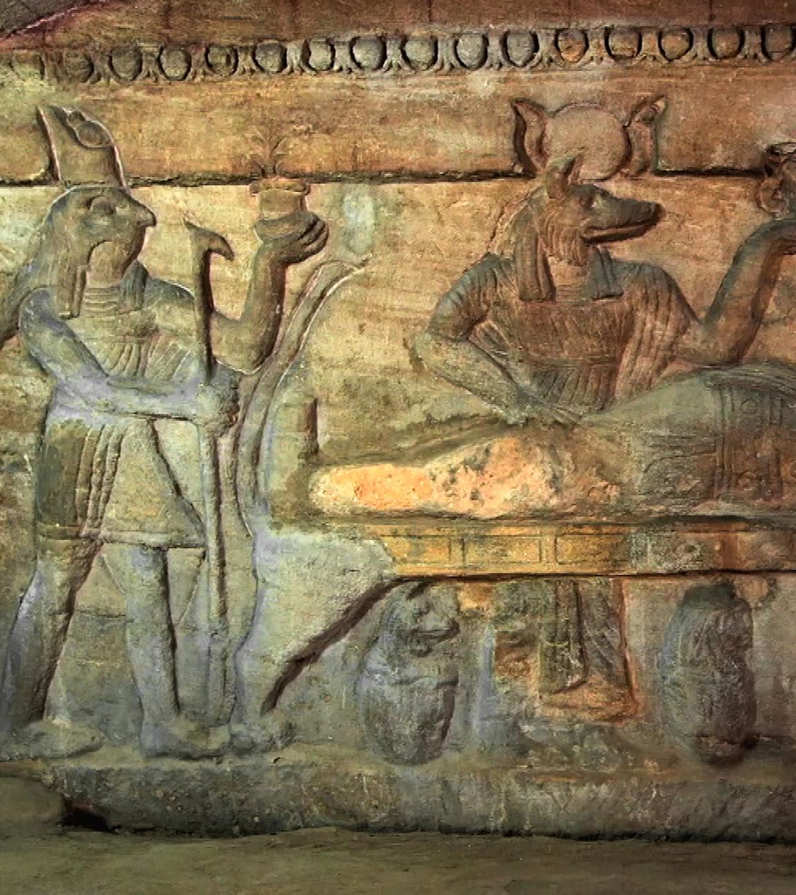 Wandrelief, bei dem in der Mitte ein Leichnam aufgebahrt liegt. Am Fußende des Toten steht Horus, der Schutzgott des Königs, mit Falkenkopf. Mittig steht der Totengott Anubis mit Schakalkopf. Beide Götter halten ein Gefäß in ihrer linken Hand.