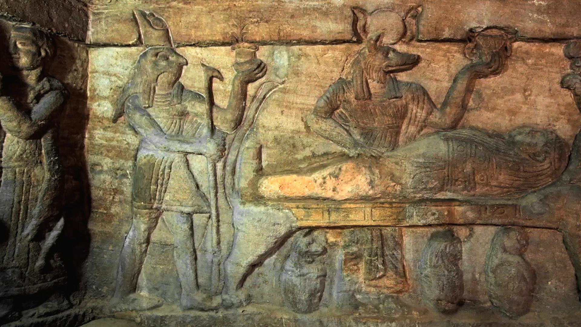 Wandrelief, bei dem in der Mitte ein Leichnam aufgebahrt liegt. Am Fußende des Toten steht Horus, der Schutzgott des Königs, mit Falkenkopf. Mittig steht der Totengott Anubis mit Schakalkopf. Beide Götter halten ein Gefäß in ihrer linken Hand.
