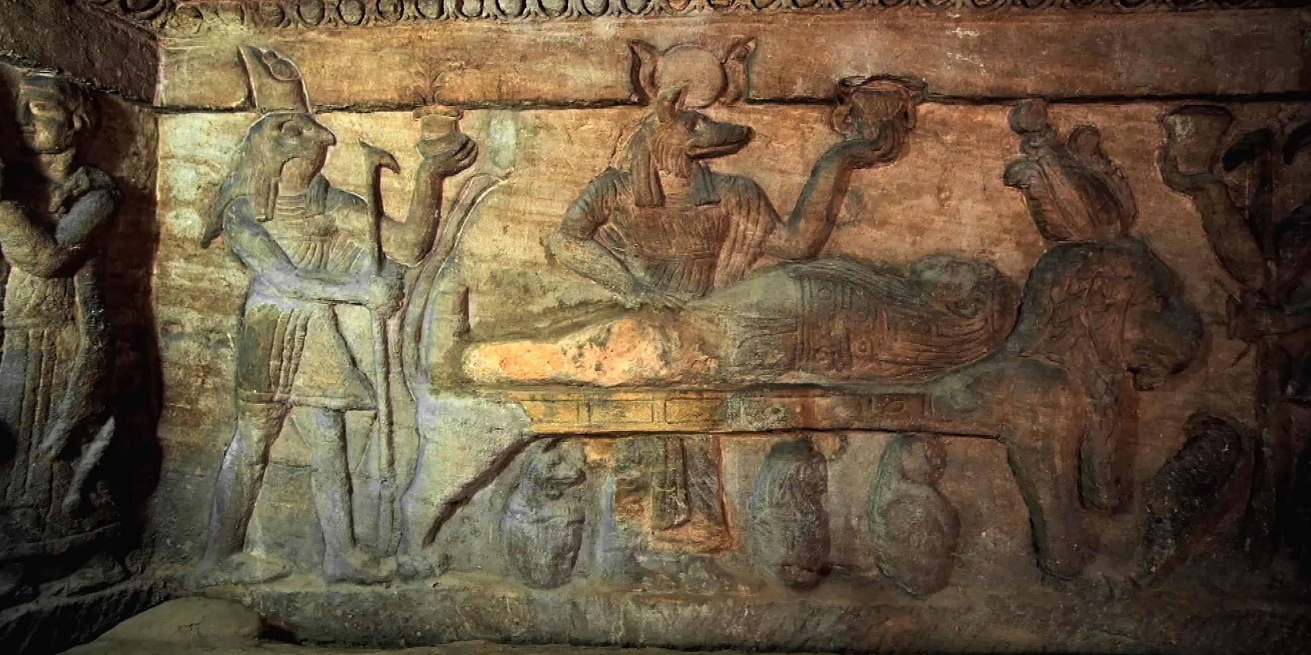 Wandrelief, bei dem in der Mitte ein Leichnam aufgebahrt liegt. Am Fußende des Toten steht Horus, der Schutzgott des Königs, mit Falkenkopf. Mittig steht der Totengott Anubis mit Schakalkopf. Beide Götter halten ein Gefäß in ihrer linken Hand.