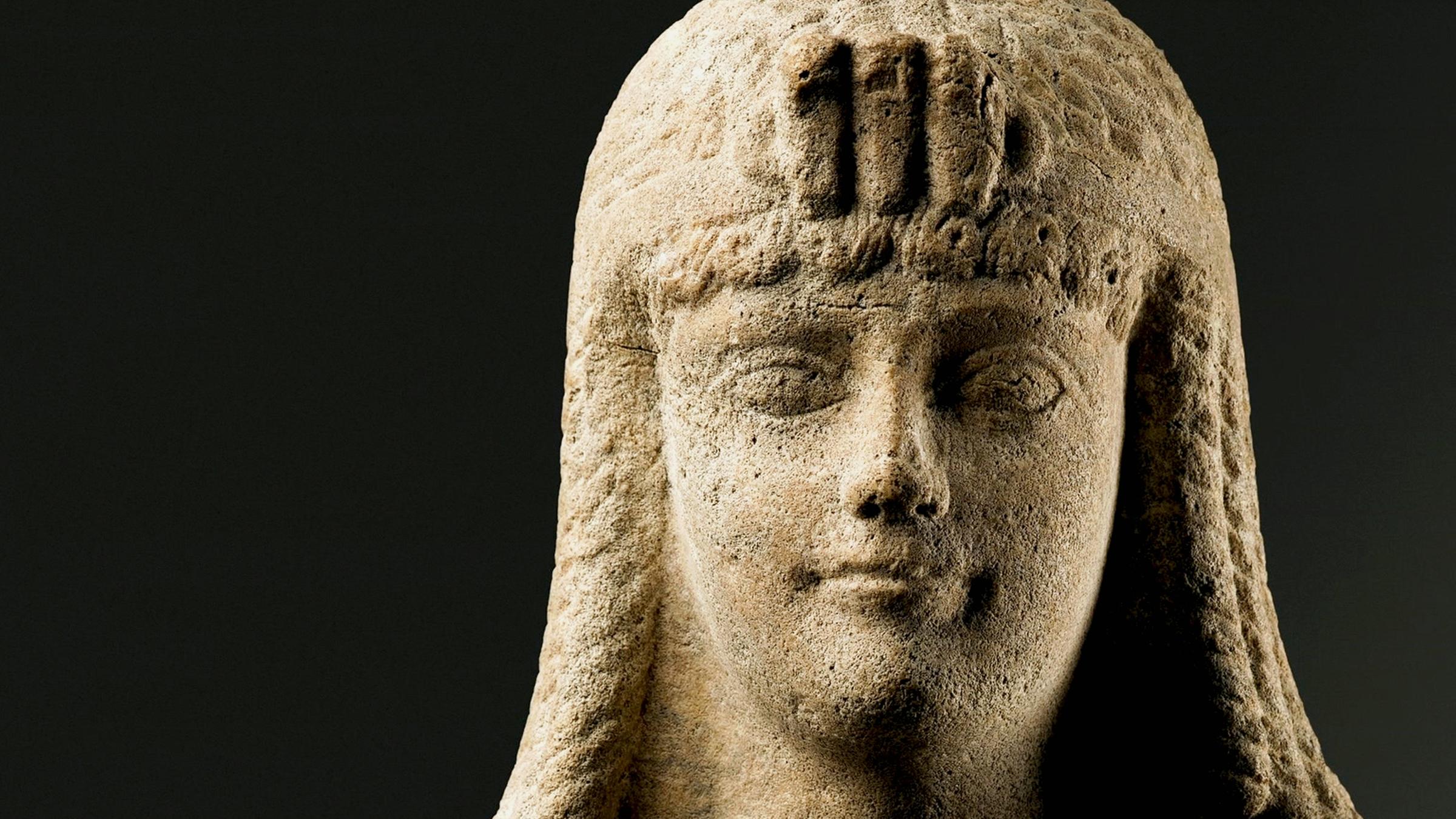 Steinerne Statue einer Frau mit langen, geflochtenen Haaren und einer Haube mit Verzierungen auf der Stirn.