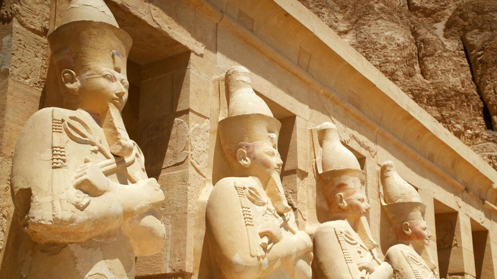 Vier Statuen männlicher Pharaonen stehen vor einem Tempel. Sie tragen Kronen, Kinnbärte, haben verschränkte Arme und halten Krummstab und Geißel in ihren Händen.