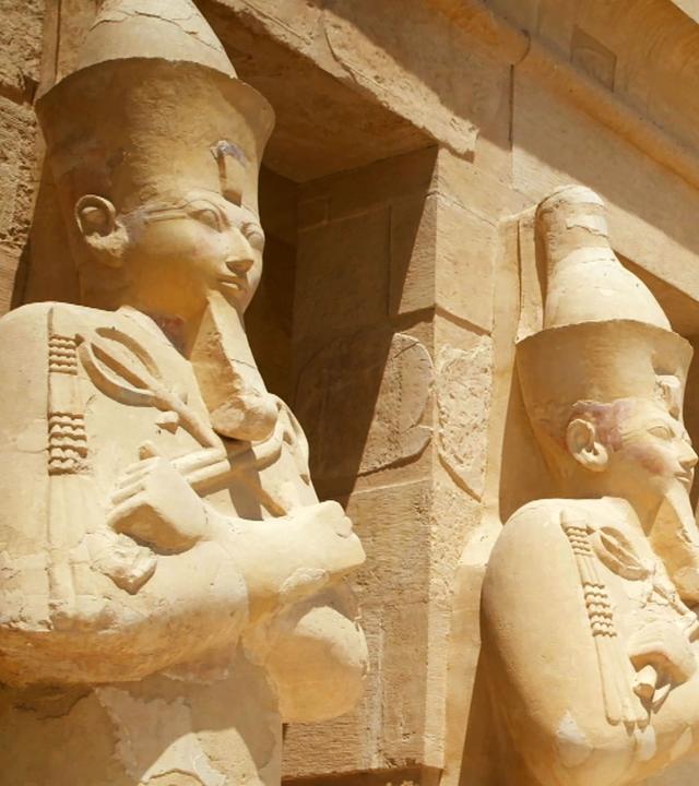 Vier Statuen männlicher Pharaonen stehen vor einem Tempel. Sie tragen Kronen, Kinnbärte, haben verschränkte Arme und halten Krummstab und Geißel in ihren Händen.