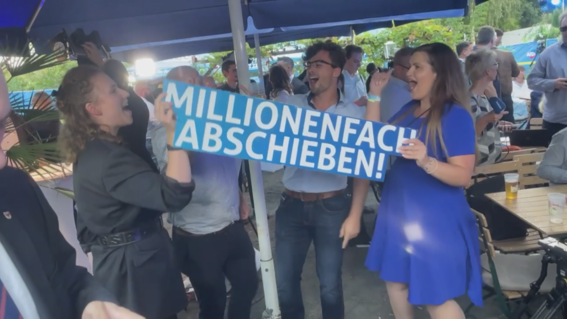 AfD-Mitglieder stehen unter einem blauen Sonnenschirm, singen und halten dabei ein Schild mit der Aufschrift: "millionenfach abschieben"