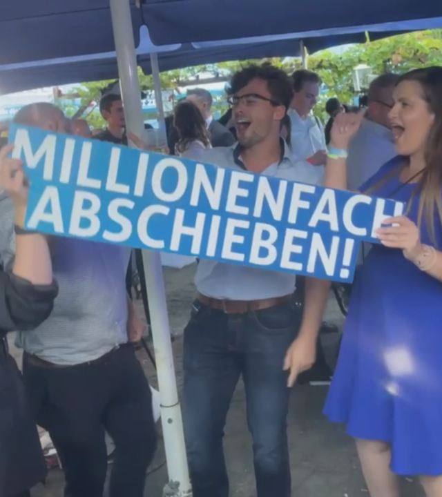 AfD-Mitglieder stehen unter einem blauen Sonnenschirm, singen und halten dabei ein Schild mit der Aufschrift: "millionenfach abschieben"