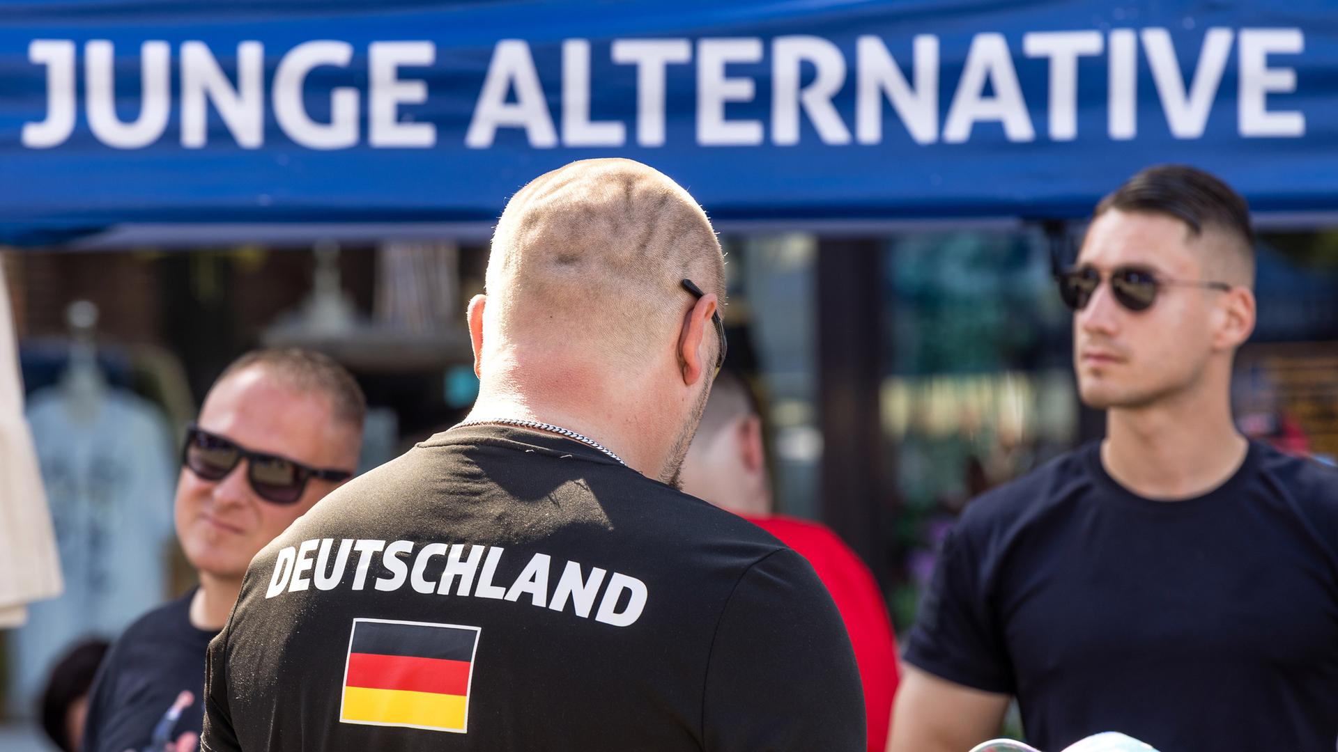 Rückansicht eines kahl rasierten Mannes mit der Aufschrift Deutschland auf seinem T-Shirt vor einem Stand der Jungen Alternativen.