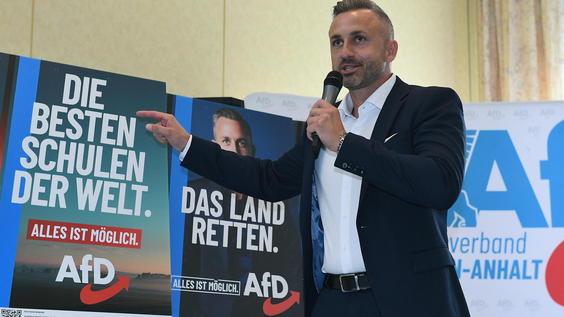 Ulrich Siegmund hält ein Mikrofon in de Hand und zeigt auf Wahlplakate der AfD mit der Aufschrift "Die besten Schulen der Welt" und "Das Land retten".