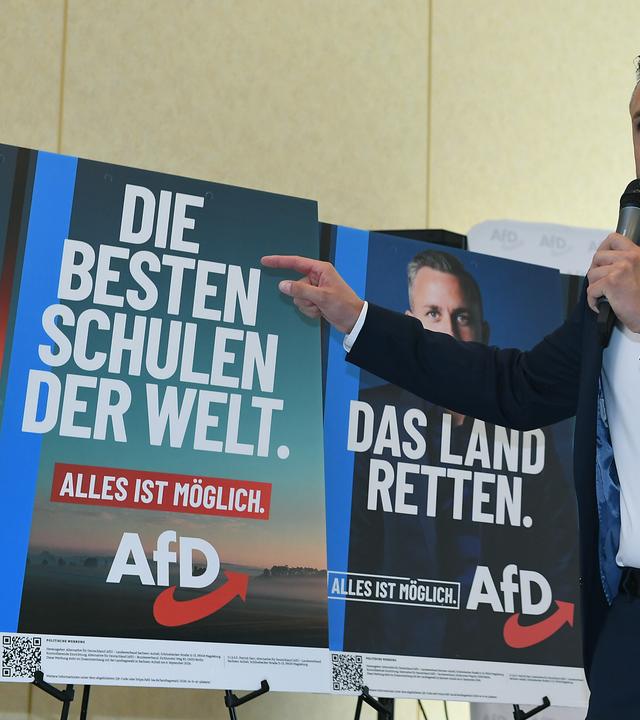 Ulrich Siegmund hält ein Mikrofon in de Hand und zeigt auf Wahlplakate der AfD mit der Aufschrift "Die besten Schulen der Welt" und "Das Land retten".