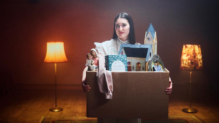 Gloria steht zusammen mit ihrem Umzugskarton im spärlich beleuchteten Wohnzimmer. Im Karton befindet sich eine Playmobil-Kirche, eine Bibel, eine Marienfigur und ein Foto mit ihrem Ehemann.