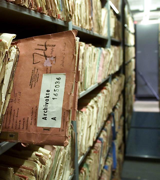 Dicht gefüllte Archivregale mit alten Akten. Im Vordergrund ragt eine abgegriffene, rötlich-braune Akte aus dem Regal.