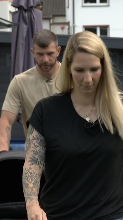 Zu sehen ist im Vordergrund eine blondhaarige Frau; hinter ihr läuft ein Mann mit Kinderwagen.