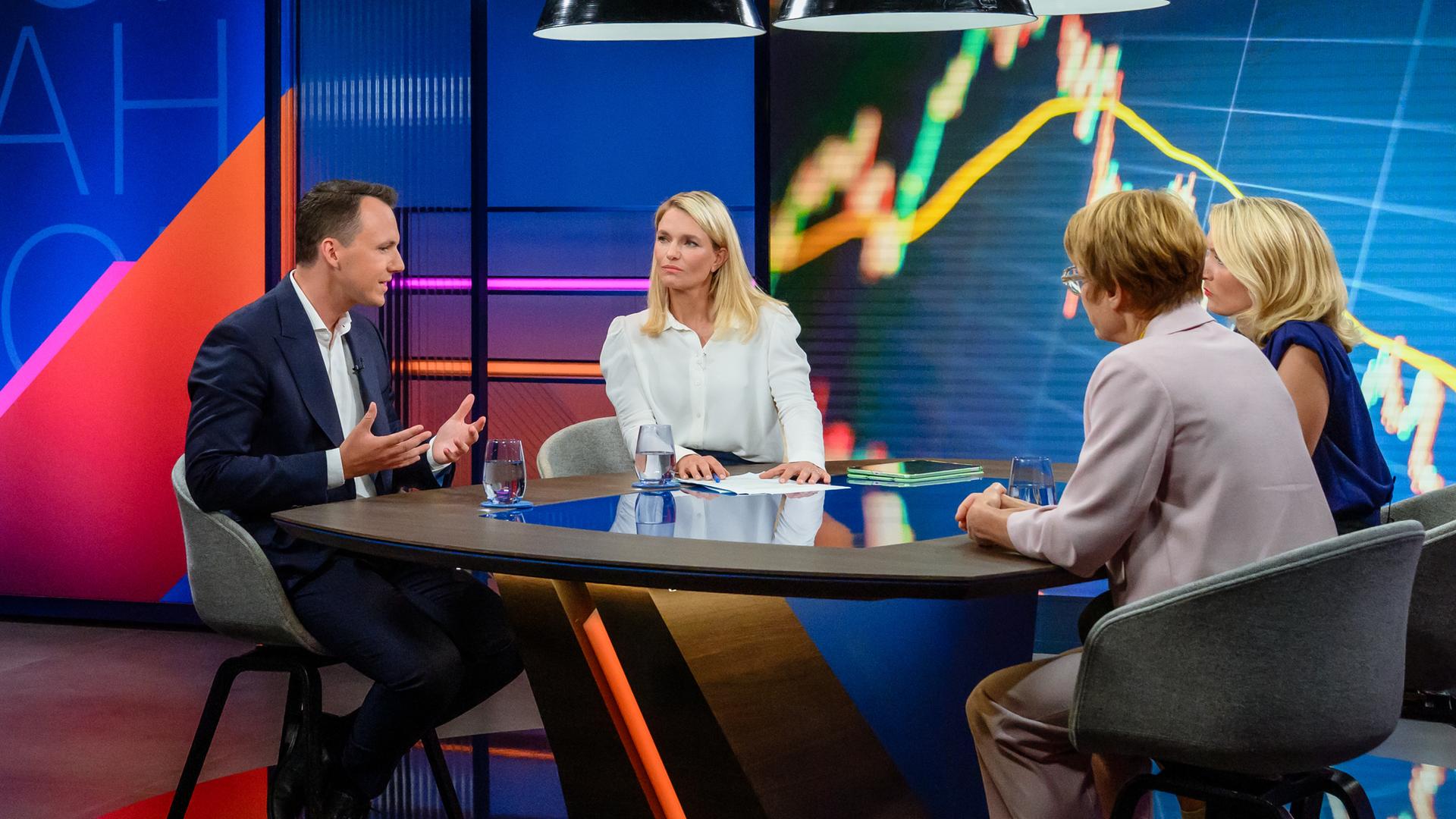 Sarah Tacke am Tisch mit ihren Gästen im Studio, an den Wänden sind bunte Grafiken zu sehen.