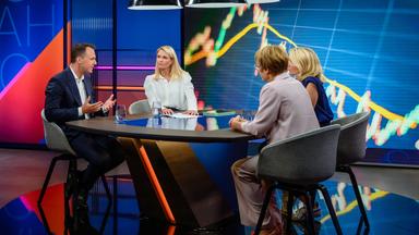 Sarah Tacke am Tisch mit ihren Gästen im Studio, an den Wänden sind bunte Grafiken zu sehen.