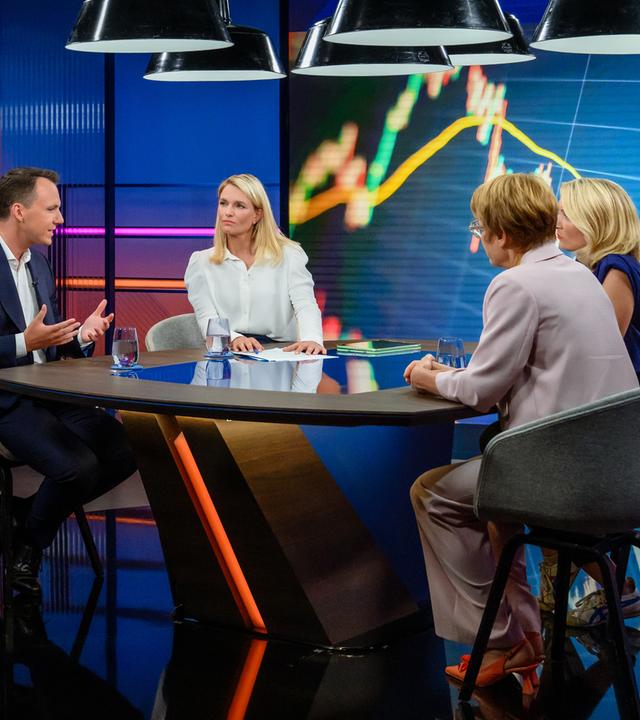 Sarah Tacke am Tisch mit ihren Gästen im Studio, an den Wänden sind bunte Grafiken zu sehen.