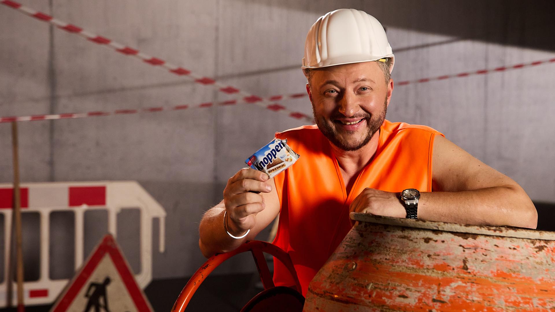Sebastian Lege steht als Bauarbeiter verkleidet in einem Baustellen-Set und hält lächelnd ein "Knoppers" in der Hand.
