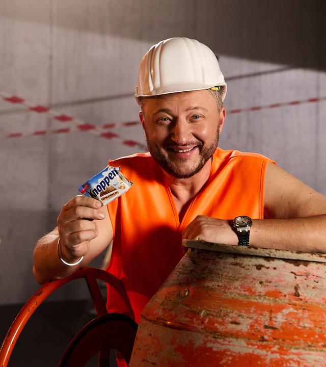 Sebastian Lege steht als Bauarbeiter verkleidet in einem Baustellen-Set und hält lächelnd ein "Knoppers" in der Hand.