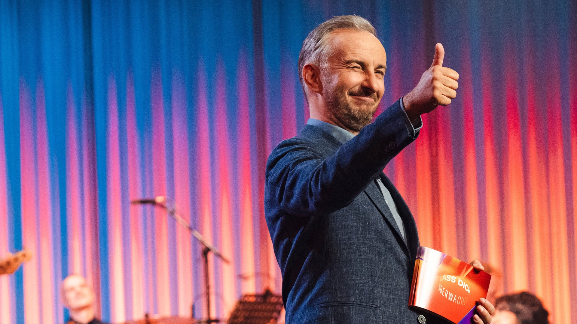 Jan Böhmermann sitzt gemeinsam mit dem Maskottchen Leaky im Publikum. Er lacht und klatscht.