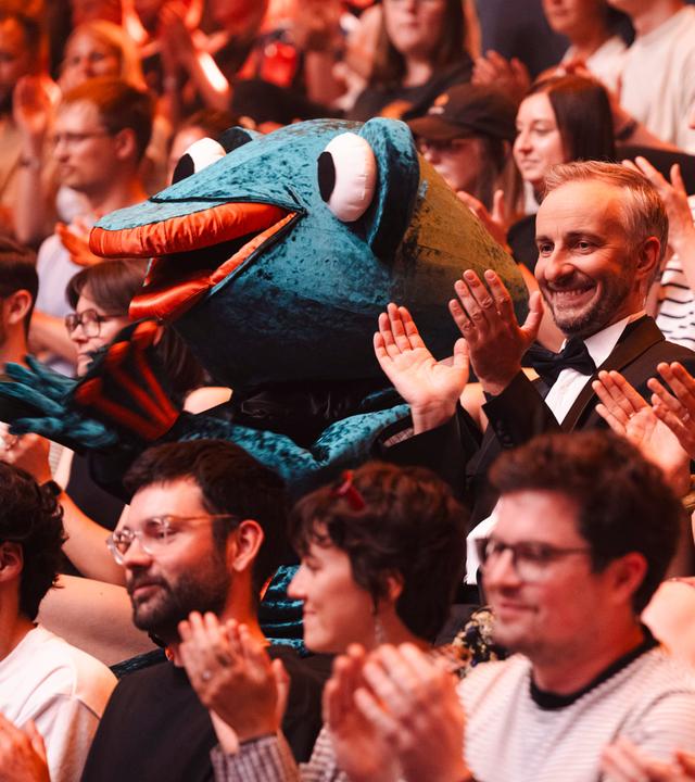 Jan Böhmermann sitzt gemeinsam mit dem Maskottchen Leaky im Publikum. Er lacht und klatscht.