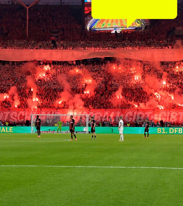 Blick auf das Fußballstadion mit roter Fankurve im Hintergrund und Pyrotechnik