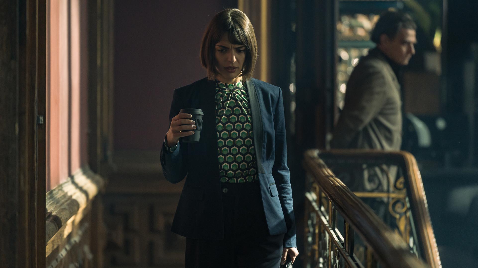 Mit einer Kaffee-to-go Tasse in der einen, einer Tasche in der anderen Hand schreitet die elegante Julia Bremora (Deniz Orta) durch die holzgetäferten Gänge des Verlags. Im Hintergrund in der Unschärfe ist Waldemar Bär (Martin Feifel) zu sehen, der in eine andere Richtung geht.