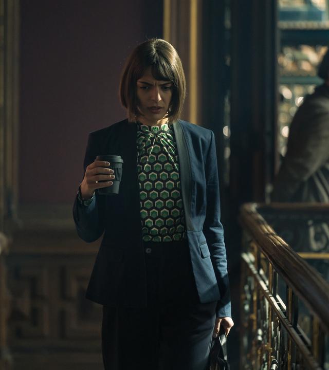 Mit einer Kaffee-to-go Tasse in der einen, einer Tasche in der anderen Hand schreitet die elegante Julia Bremora (Deniz Orta) durch die holzgetäferten Gänge des Verlags. Im Hintergrund in der Unschärfe ist Waldemar Bär (Martin Feifel) zu sehen, der in eine andere Richtung geht.