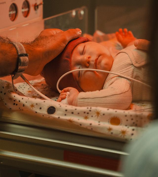 "PUSH - Urvertrauen": Ida (Johanna Reinders) und ihr Freund Giovanni (Hassan Kello) berühren vorsichtig ihr Baby, das in der Klinik auf einem Babybett liegt. Idas Hand liegt auf seinem Bauch, Giovannis Hand auf seinem Kopf.