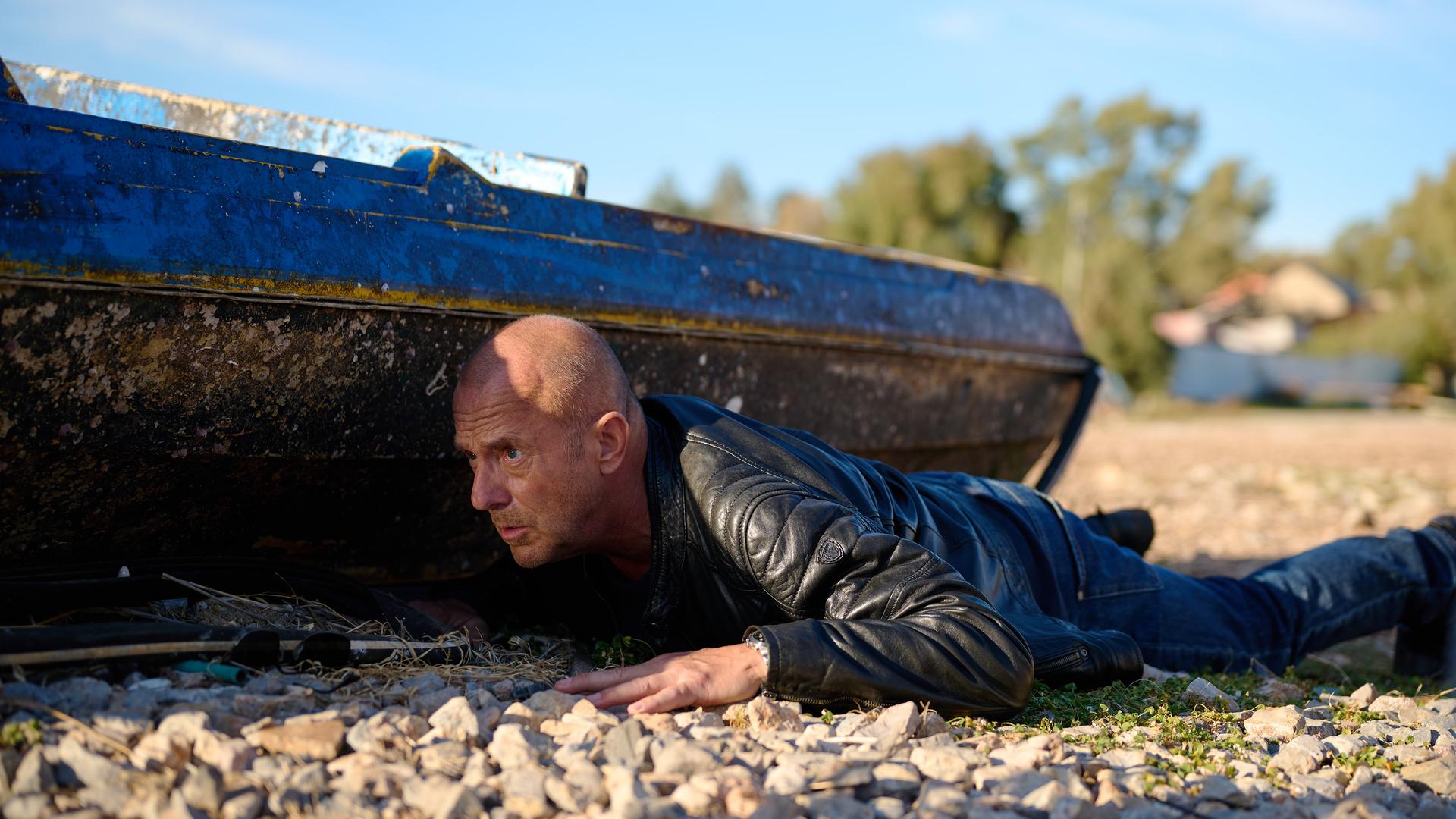Ingo Thiel (Heino Ferch) liegt auf einem Schotterboden. Er versteckt sich hinter einem alten Boot und beobachtet angespannt etwas.