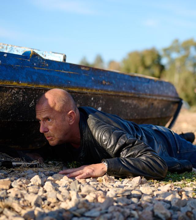 Ingo Thiel (Heino Ferch) liegt auf einem Schotterboden. Er versteckt sich hinter einem alten Boot und beobachtet angespannt etwas.