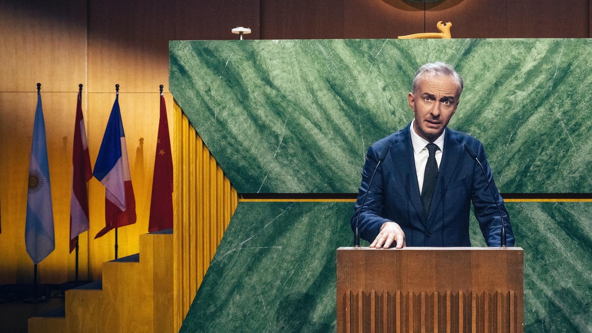 Jan Böhmermann steht am Rednerpult auf der Bühne der "International Comedy Union". Hinter ihm ist ein Podium für den Vorsitz der "International Comedy Union" zu sehen, rechts und links davon verschiedener Länderflaggen. An der Wand ganz im Hintergrund ist oben das Logo der "International Comedy Union" - zwei Clownshände im Handschlag mit einem Globus und einem Lorbeerkranz - zu sehen.