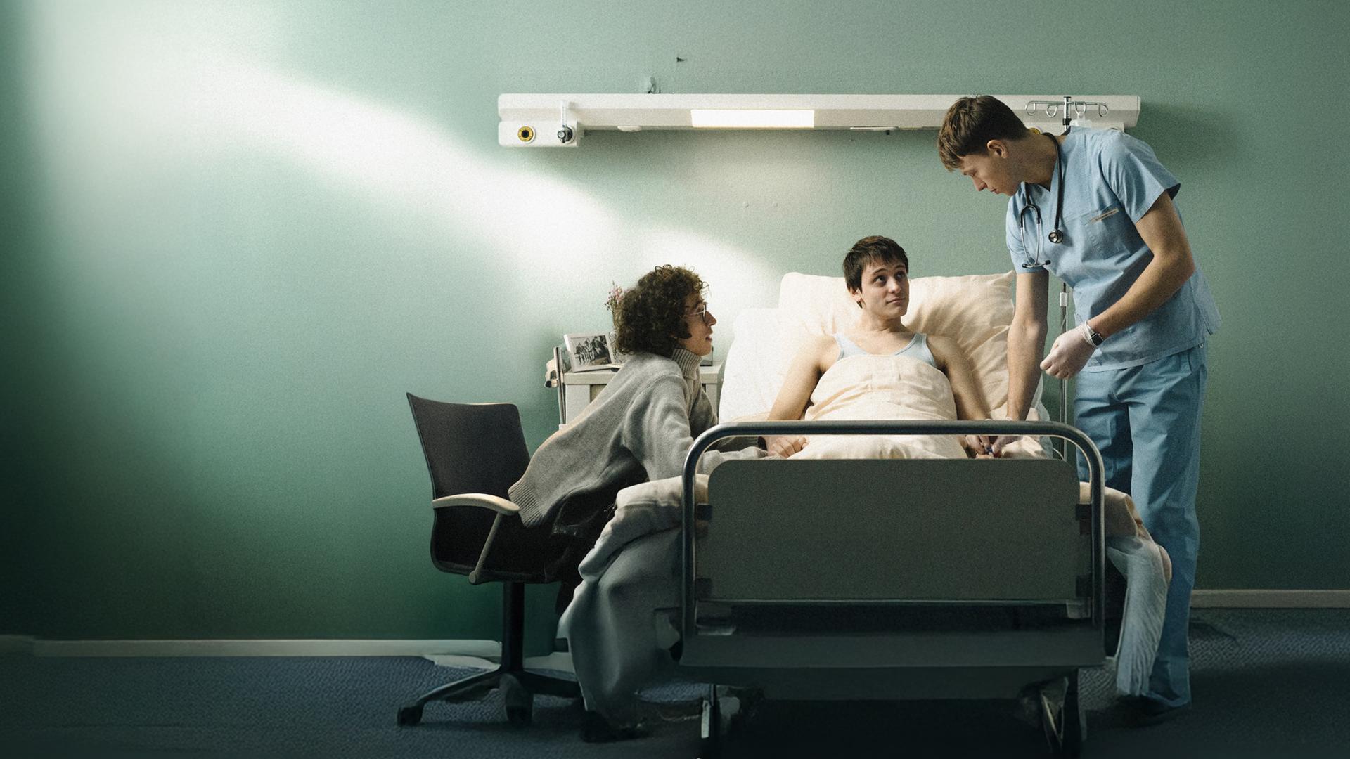 Heiko (Evangelos Tsarkowistas) am Bett - er hält Heikos Hand,. Auf der anderen Seite steht der Krankenpfleger Bernhard (Clemens Bobke).