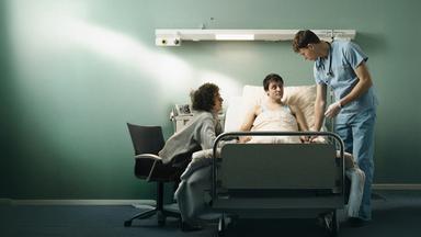 Dirk (Evangelos Tsarkowistas) am Bett - er hält Heikos Hand,. Auf der anderen Seite steht der Krankenpfleger Bernhard (Clemens Bobke).