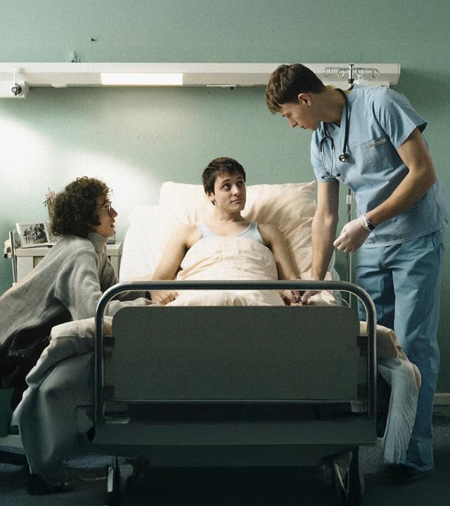 Heiko (Evangelos Tsarkowistas) am Bett - er hält Heikos Hand,. Auf der anderen Seite steht der Krankenpfleger Bernhard (Clemens Bobke).