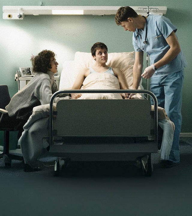 Dirk (Evangelos Tsarkowistas) am Bett - er hält Heikos Hand,. Auf der anderen Seite steht der Krankenpfleger Bernhard (Clemens Bobke).