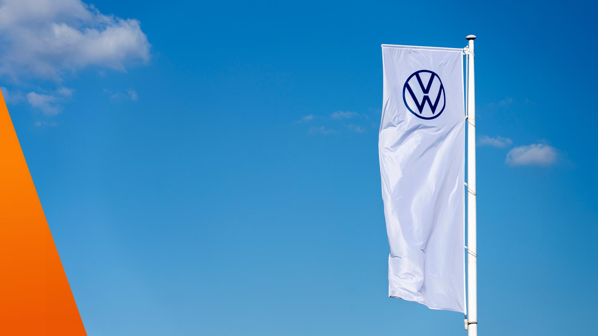 Rechts flattert eine lange weiße Flagge mit dem VW-Zeichen vor einem kräftig blauen Himmel. Links ragt eine orangefarbene, geometrische Form in das ansonsten freie Bild. Die Komposition begleitet das Thema eines möglichen europäischen Automobilherstellers nach Airbus-Vorbild, der zum Fortbestand der Marke Volkswagen beitragen soll.