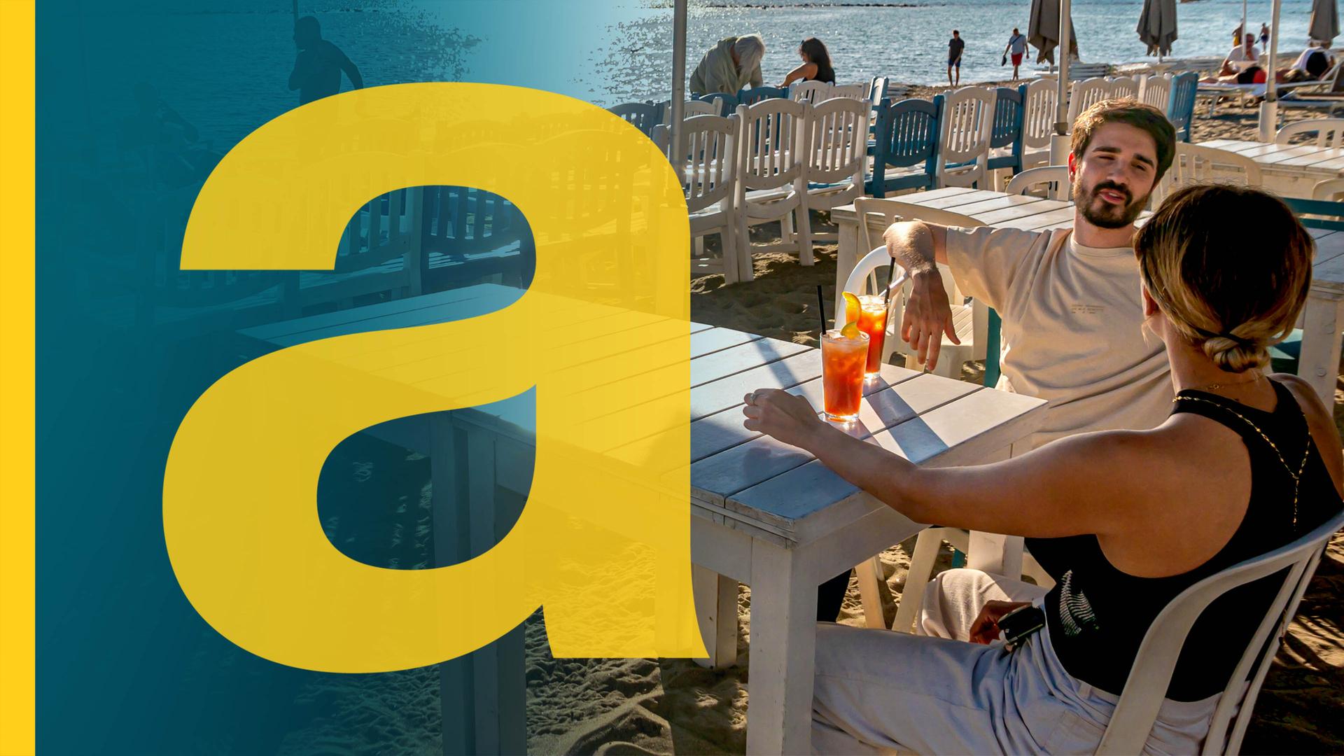 Ein Pärchen sitzt an Tisch am Strand in Italien, im Hintergrund Sonnenliegen, Meer und Touristen