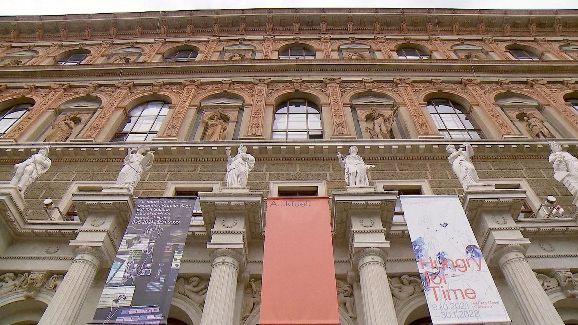 Das Bild zeigt die Fassade der Akademie der bildenden Künste in Wien. Es handelt sich um ein imposantes Gebäude mit mehreren Stockwerken, das von Säulen und Statuen geschmückt ist. Die Statuen sind auf Balustraden platziert und befinden sich auf beiden Seiten des Eingangsbereichs.   Die Fassade weist aufwendig gestaltete Elemente wie Fenster, Verzierungen und Reliefs auf, die das architektonische Gesamtbild prägen. Es sind mehrere Banner angebracht, die Informationen über aktuelle Ausstellungen enthalten.   Der Himmel ist bewölkt, was dem Szenario eine gedämpfte Atmosphäre verleiht. Die Akademie gilt als eine der ältesten Kunstakademien in Europa und hat zahlreiche berühmte Künstler hervorgebracht.