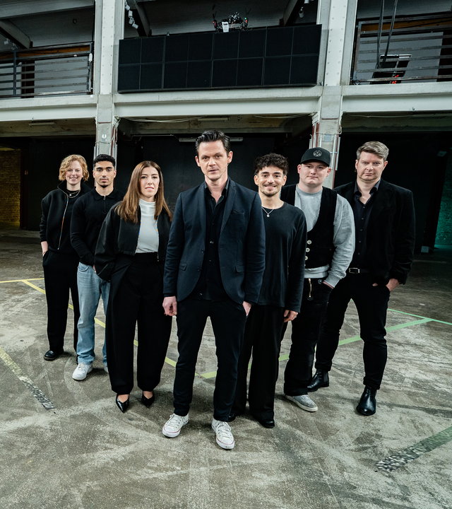 Es stehen sieben Menschen auf einem Boden, der ein schachbrettartiges Muster hat. Die sieben Menschen stehen in einer Reihe. Es sind zwei Frauen und fünf Männer. Alle blicken in die Richtung des Betrachters.